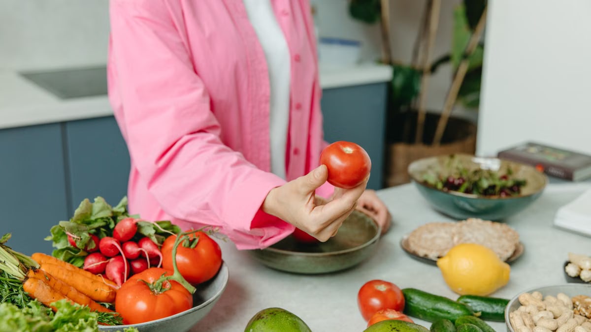 Person in pink overshirt holding a fresh tomato with various organic vegetables on a countertop.